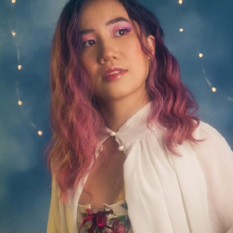 Photo of Tiffany shows a Vietnamese person with long wavy black to pink gradient hair, pink eyeshadow, and wearing a collared white cape and a white corset with red flowers. They are looking into the distance, and the background is a light blue with fairy lights.