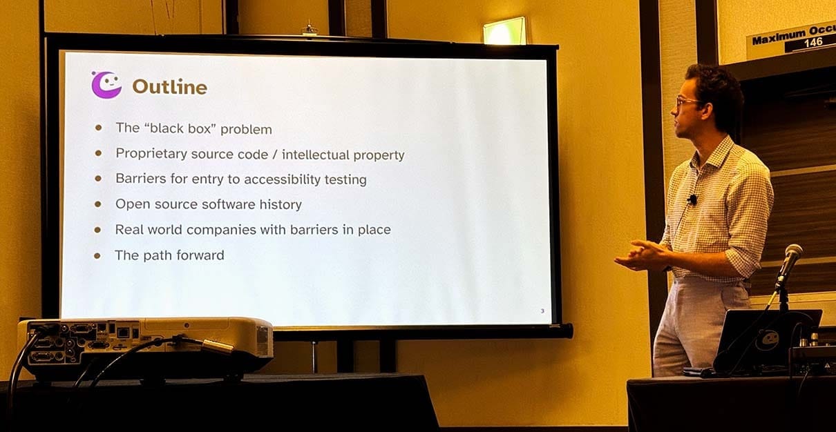 Nicholas Cook, software engineer, stands in front of a screen showing an outline of his CSUN talk. The bullets read: The “black box” problem, Proprietary source code / intellectual property, Barriers for entry to accessibility testing, Open source software history, Real world companies with barriers in place, and The path forward